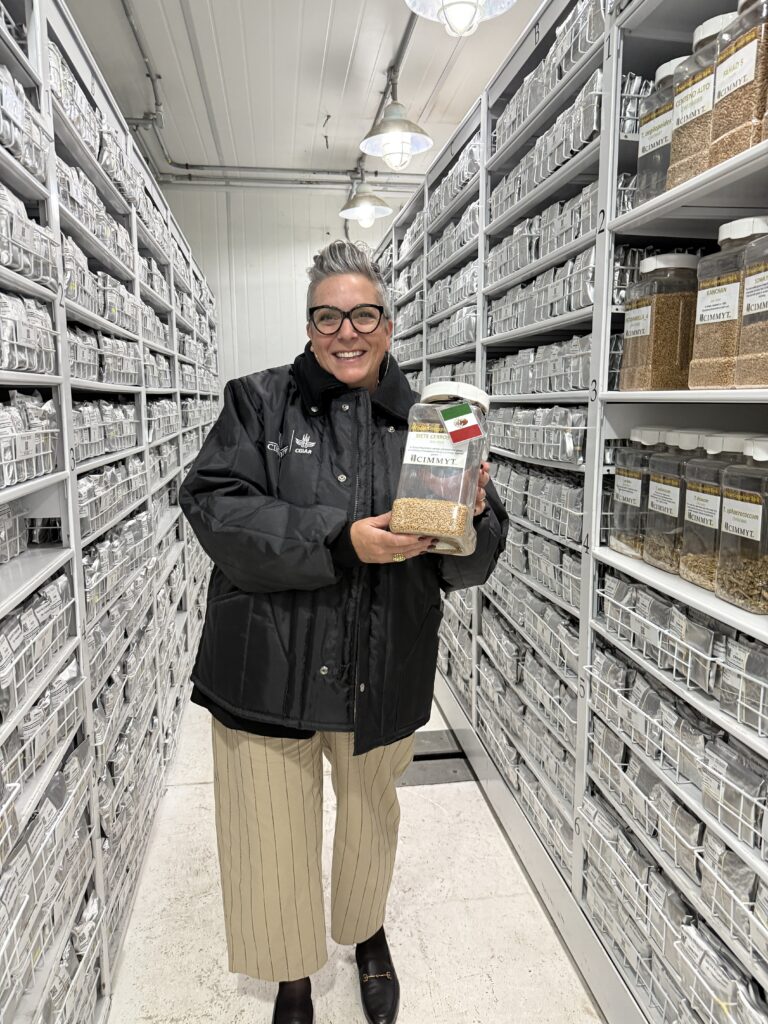 A stylish woman with short, gray hair and glasses smiles brightly at the camera, holding a large jar of grains. She is dressed in a black quilted jacket, tan pinstriped pants, and black loafers. She stands in a long, narrow storage room with rows of shelves on either side, filled with countless jars of what appear to be seeds or grains. The room is well-lit by overhead industrial lights, and the overall impression is one of organized research or collection.