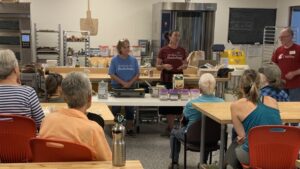Two women and a man are presenting at a table in a modern kitchen or classroom to a seated audience. The table holds several packages and bowls, including a box of buckwheat. The women are wearing t-shirts that say "Buckwheat."