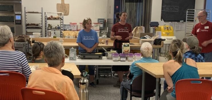 Two women and a man are presenting at a table in a modern kitchen or classroom to a seated audience. The table holds several packages and bowls, including a box of buckwheat. The women are wearing t-shirts that say "Buckwheat."