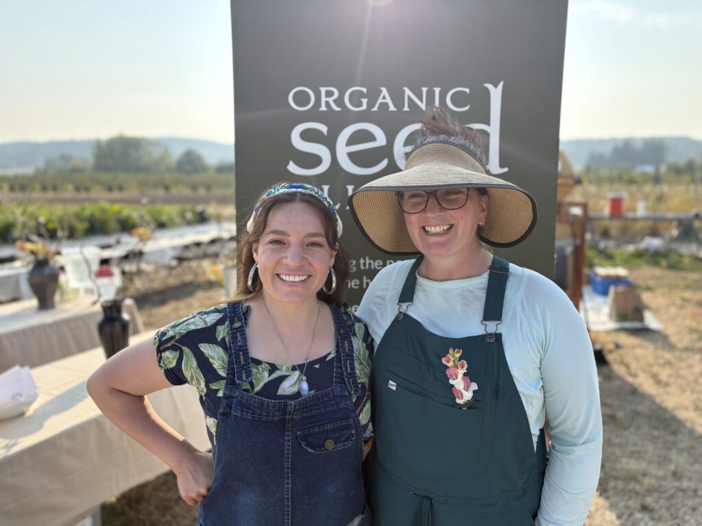 Two smiling women stand in a field with a large green banner behind them that reads "ORGANIC seed." The woman on the left has a blue headband and a floral shirt with overalls. The woman on the right wears a wide-brimmed straw hat and a light blue shirt under a green apron.