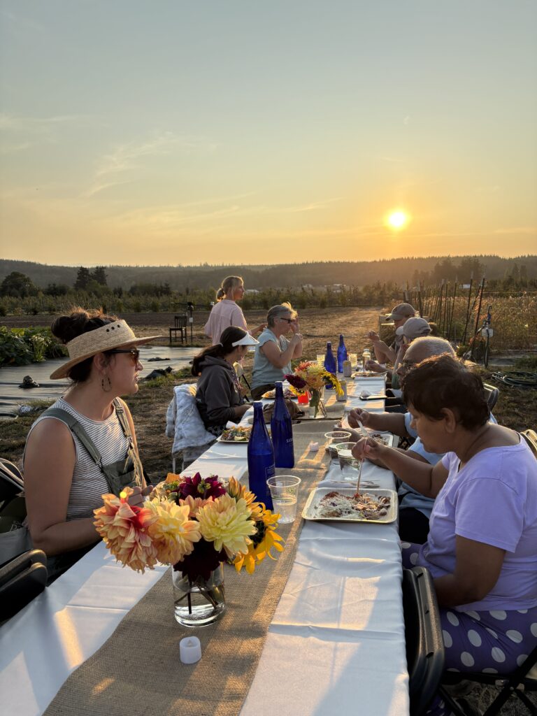 A group of people sits at a long table covered with a white tablecloth in a field at sunset. The table is set with blue water bottles, flowers in vases, and plates of food. A woman in a striped shirt and a straw hat is in the foreground, facing another person.