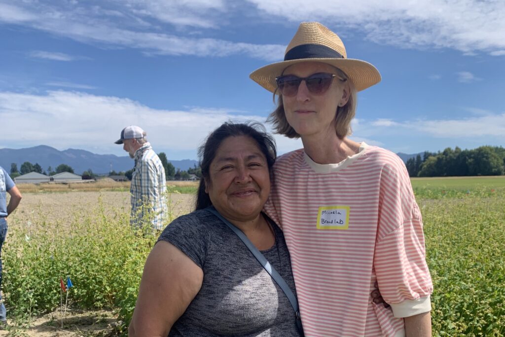 Two women, Micaela and Nelida, stand side-by-side in a field under a bright sky, smiling at the camera.