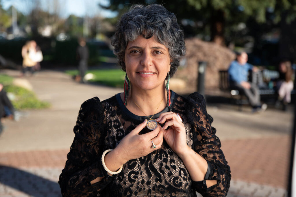 A headshot of Vivien Sansour, the founder of the Palestine Heirloom Seed Library and a 2025 Organic Seed Growers Conference Keynote Speaker. She is wearing a black lace-patterned dress and large, beaded earrings, and is holding a necklace with a round pendant. The background is a park with blurry trees and people. The photo was taken by Matthew Stover of Delta Creative Agency.