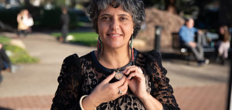 A headshot of Vivien Sansour, the founder of the Palestine Heirloom Seed Library and a 2025 Organic Seed Growers Conference Keynote Speaker. She is wearing a black lace-patterned dress and large, beaded earrings, and is holding a necklace with a round pendant. The background is a park with blurry trees and people. The photo was taken by Matthew Stover of Delta Creative Agency.