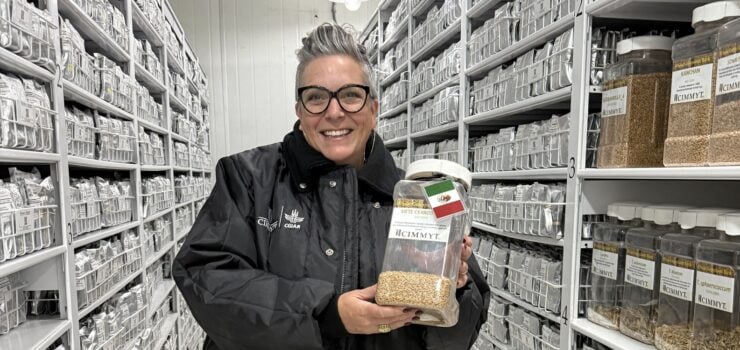 A stylish woman with short, gray hair and glasses smiles brightly at the camera, holding a large jar of grains. She is dressed in a black quilted jacket, tan pinstriped pants, and black loafers. She stands in a long, narrow storage room with rows of shelves on either side, filled with countless jars of what appear to be seeds or grains. The room is well-lit by overhead industrial lights, and the overall impression is one of organized research or collection.