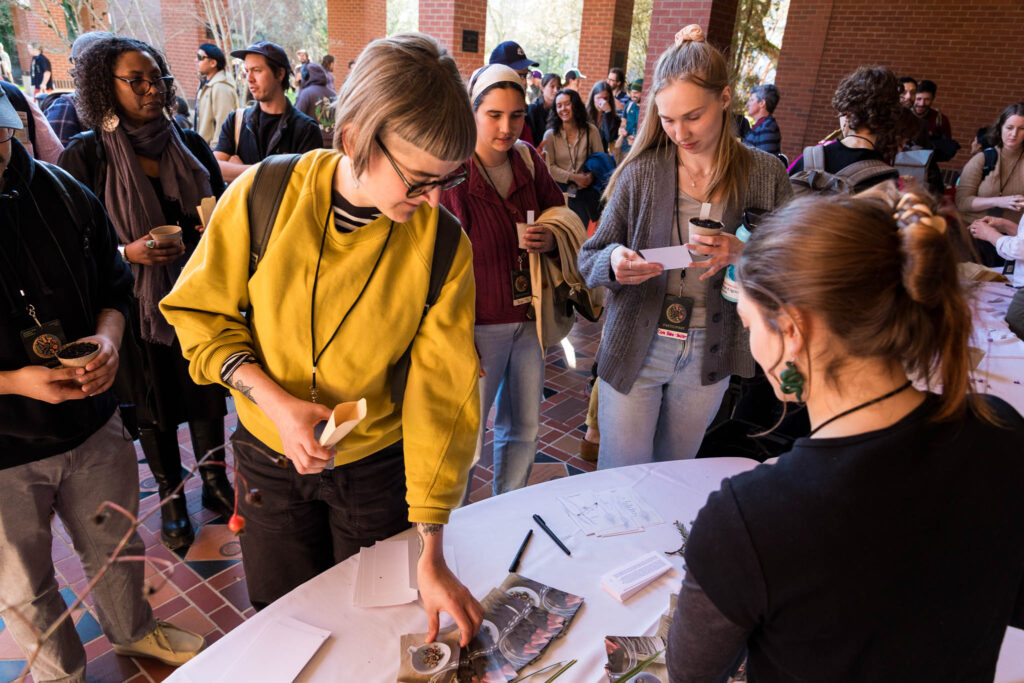 A group of people standing around a white table at an indoor event. A person in a yellow sweater is on the left, looking down at the table. A person with a headband and a young woman with long blonde hair stand behind them. The person on the right, wearing a black shirt, is seated at the table, helping attendees.