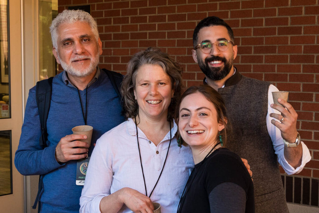 A group of four people standing together and smiling in front of a brick building. From left to right: a man with gray hair and a blue shirt, a woman with graying hair in a light-colored shirt, a young woman with a long dark ponytail in a black shirt, and a man with a beard and glasses wearing a patterned vest. They are all holding small paper cups.