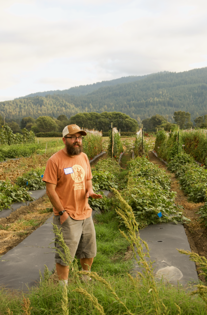 Organic farmer giving a field tour, standing among rows of vine crops with mountains in the background. Organic seed stewardship.